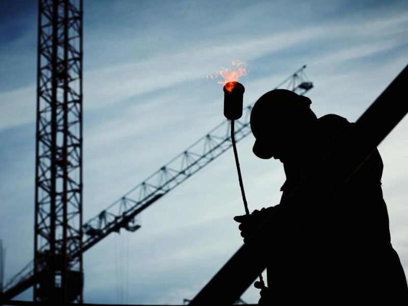 Silhouette of a construction worker using a blowtorch at a building site against a crane-filled skyline.